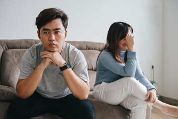 Fototapeta premium Unhappy Asian spouses sitting on couch back to back sulking after quarrel at home. Family conflict and domestic problem