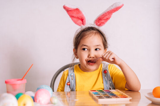 Little girl wearing bunny ears on her head eats chocolate from spoon.