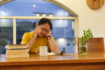 A middle-aged woman reading a book; her hands on her neck; a stack of books, a cup and a vase nearby - obrazy, fototapety, plakaty