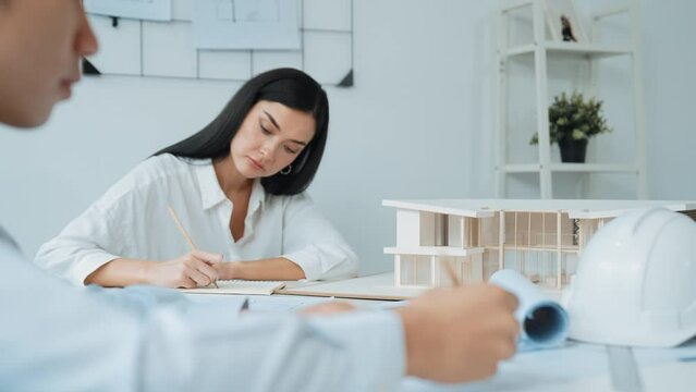 Professional architect engineer drawing blueprint and working together with young beautiful caucasian coworker on meeting table with safety helmet, house model, blueprint scatter around. Immaculate.