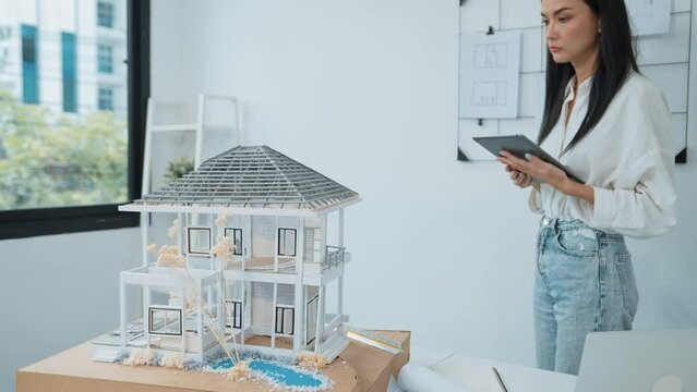 Beautiful caucasian architect focus on using tablet to check house model on meeting table with blueprint, house model and architectural document scatter around at meeting room. Immaculate.