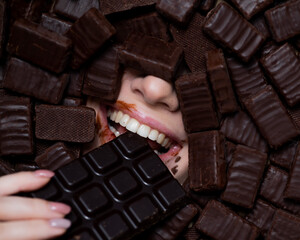 The face of a caucasian woman surrounded by chocolates. The girl eats a bar of chocolate.