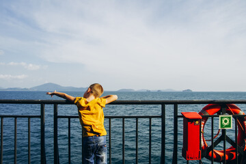 A boy in a yellow T-shirt stands on the deck of the ferry and looks at the sea. The child looks into the distance