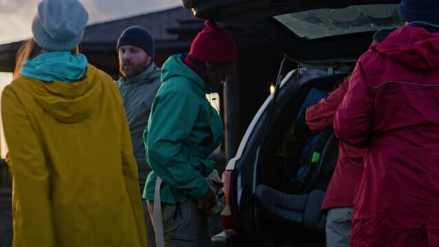 Group of diverse hikers unpack luggage and backpacks from trunk of car, settle into rented house in mountains. Tourist family or friends during vacation trip. Tourism and active leisure. Slow motion.