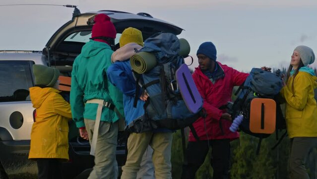 African American hiker takes backpacks from trunk and gives to his friends. Diverse group of tourist arrive to rental house on car. Hiking buddies during trip or trek to mountains on their vacation.
