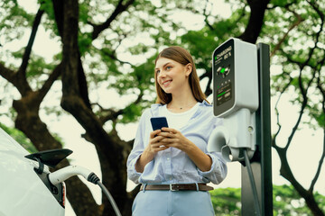 Young woman using smartphone online banking application to pay for electric car battery charging...