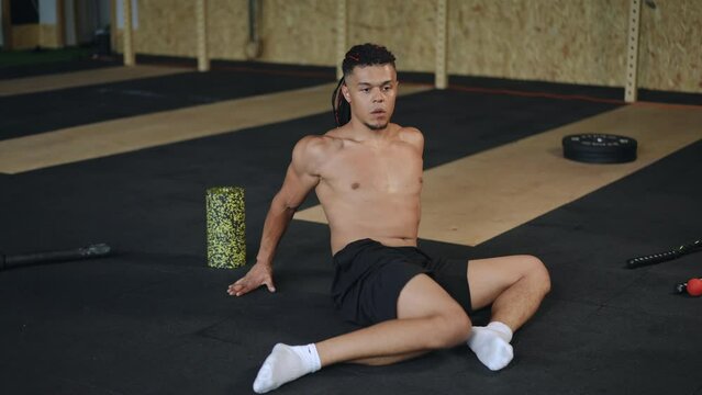 A mixed race man with dreadlocks on his head does a dynamic hip stretching exercise while sitting on the floor in the gym