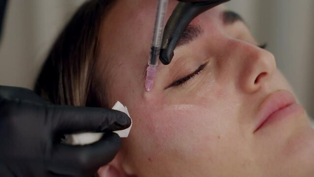 Close up Botox injection. doctor's hands in gloves doing beauty injections of botox on young woman patient cheek laying on bed and wipe gently with a cotton ball after injection in the beauty clinic.