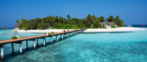 Wooden bridge over the sea leading to an island with sandy beaches and a green forest. Palm trees.