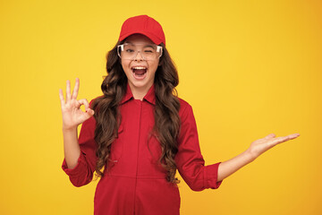 Excited teenager girl. Worker teenager child wearing overalls red, cap and protect glasses. Studio shot portrait isolated on yellow background. Pointing and showing concept. - obrazy, fototapety, plakaty