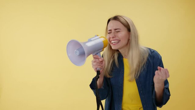 Nervous angry female holding white loudspeaker and shouting in it over isolated yellow studio background. Emotional caucasian woman gesturing with hand and raising voice to be hearding indoors.