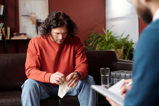 Young man suffering depression holding tissue sitting on couch with head lowered at therapy session
