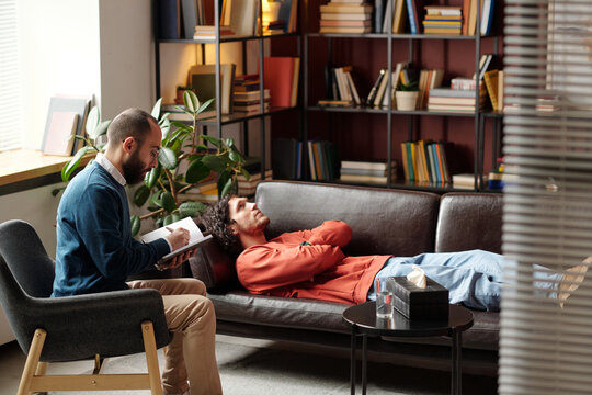 Wide shot of Middle Eastern psychologist taking notes while listening to his Caucasian patient during therapy session in modern office