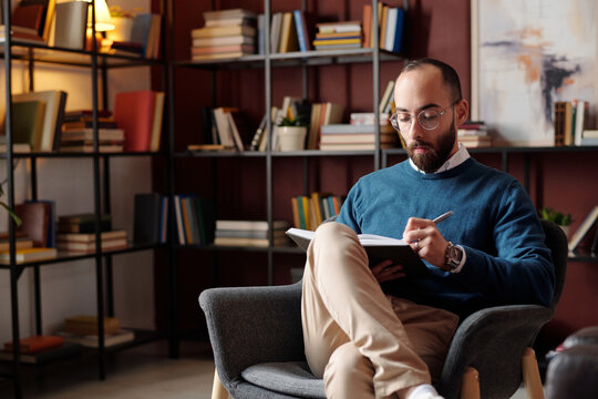 Portrait of young Middle Eastern psychologist sitting on chair in his office taking notes in notebook, copy space