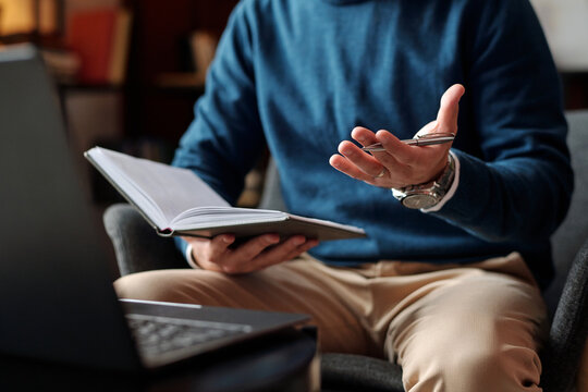 Unrecognizable psychologist holding pen and notebook sitting in front of laptop having video call with patient or colleague