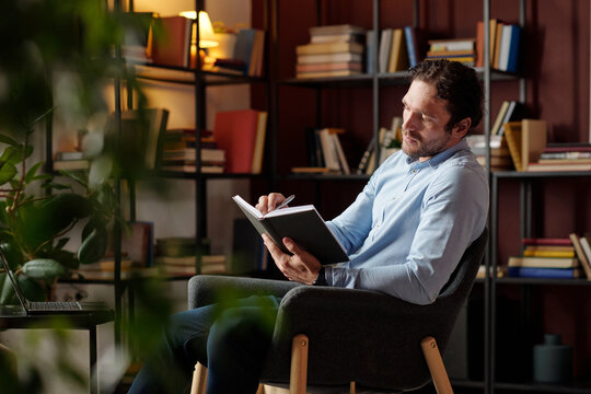 Medium long shot of young Caucasian male psychotherapist sitting on chair in his office making notes in notebook