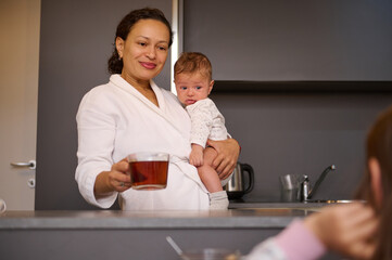 Happy smiling woman, young mother carrying her baby boy, holding a cup of hot tea while standing at kitchen counter