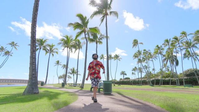 Smiling African American man with a beard wearing a floral patterned shirt and shorts walking confidently on a path lined with palm trees in a sunlit tropical park. Feeling joy. Slow Motion, 