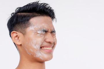 A young asian man winces in pain after getting facial scrub cream into his eyes, irritating it. Isolated on a white backdrop. - obrazy, fototapety, plakaty