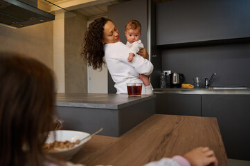 Selective focus of a young mother holding baby boy, son while older daughter sitting near bowl with delicious flakes