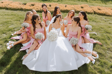 A brunette bride and her bridesmaids in identical dresses sit with the bride and rejoice with flowers in their hands on the green grass. Wedding in nature.