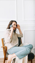 Serious millennial woman talks on the phone, sitting on a chair and looking out the window in her free time. Serious pretty millennial woman calls by phone, talks, sits on chair, looks out window.