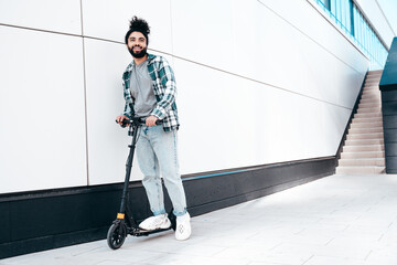 Trendy smiling bearded man in casual clothes riding electric scooter in urban background. Handsome model posing in the street near wall. Hipster guy with curly hairstyle. Cheerful and happy