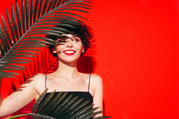 Fashion beauty portrait of young smiling brunette woman with evening stylish makeup and perfect clean skin. Sexy model with curly hair posing in studio. With red bright lips. Tropical palm leaf