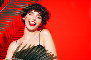 Fashion beauty portrait of young smiling brunette woman with evening stylish makeup and perfect clean skin. Sexy model with curly hair posing in studio. With red bright lips. Tropical palm leaf