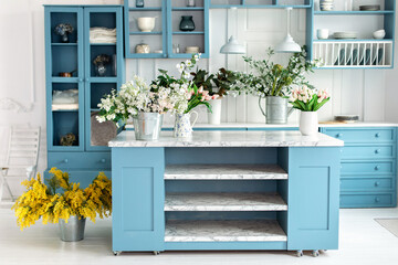 Stylish cuisine with flowers in vase. Wooden kitchen in spring decor. Cozy home decor. Kitchen island in dining room. Kitchen utensil, dishes and plate on shelves. Blue kitchen interior with furniture