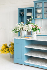 Stylish cuisine with flowers in vase. Wooden kitchen in spring decor. Cozy home decor. Kitchen island in dining room. Kitchen utensil, dishes and plate on shelves. Blue kitchen interior with furniture