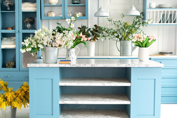Stylish cuisine with flowers in vase. Wooden kitchen in spring decor. Cozy home decor. Kitchen island in dining room. Kitchen utensil, dishes and plate on shelves. Blue kitchen interior with furniture