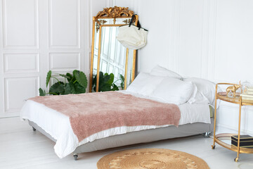 Interior white bedroom with coffee table, comfortable bed with pillows and large gold vintage mirror, copy space. Golden decorative serving table trolley in room. Cozy scandinavian decoration room