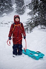 Portrait of a young European boy 7 - 9 years old in the warm winter jacket standing on the snowy winter background with sledge