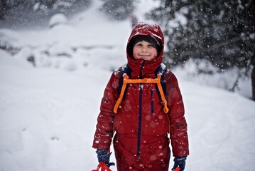 Portrait of a young European boy 7 - 9 years old in the warm winter jacket standing on the snowy winter background with sledge