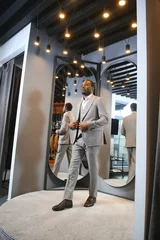 Fotobehang sale, shopping, fashion, style and people concept - african man choosing and trying jacket or suit on and looking to mirror in mall or clothing store © Serhii
