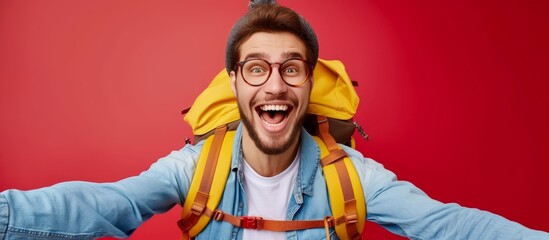 A joyful, adventurous hipster traveler in glasses, denim shirt, and a yellow backpack takes a selfie, expressing happiness and excitement on a red background.
