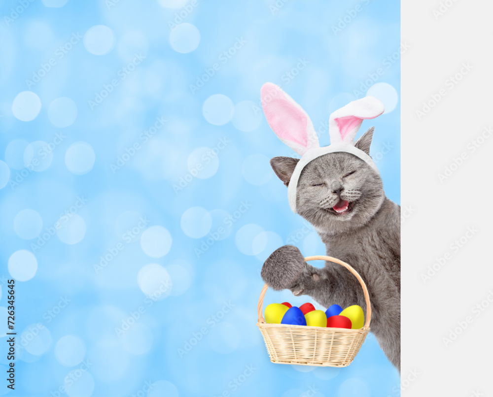 Poster happy kitten wearing easter rabbits ears holds basket with colorful easter eggs and looks from behin