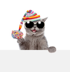 Happy cat wearing sunglasses and knitted warm woolen hat with pompon looks above empty white board. isolated on white background - obrazy, fototapety, plakaty