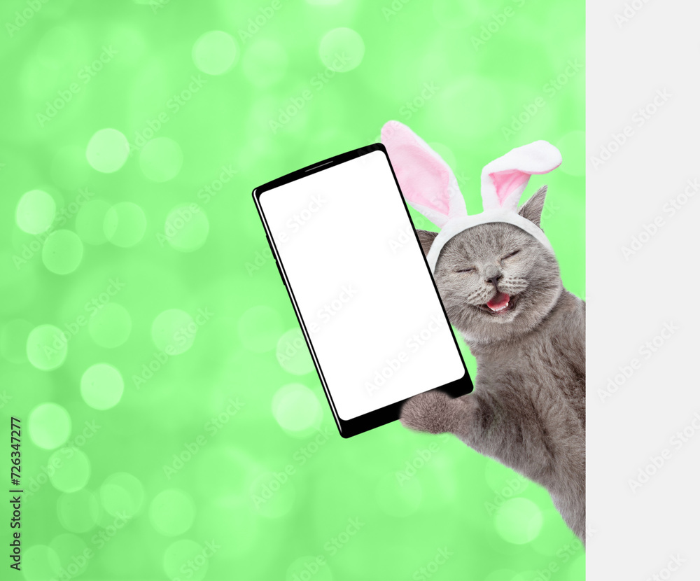 Poster happy kitten wearing easter rabbits ears holds big smartphone with white blank screen and looks from