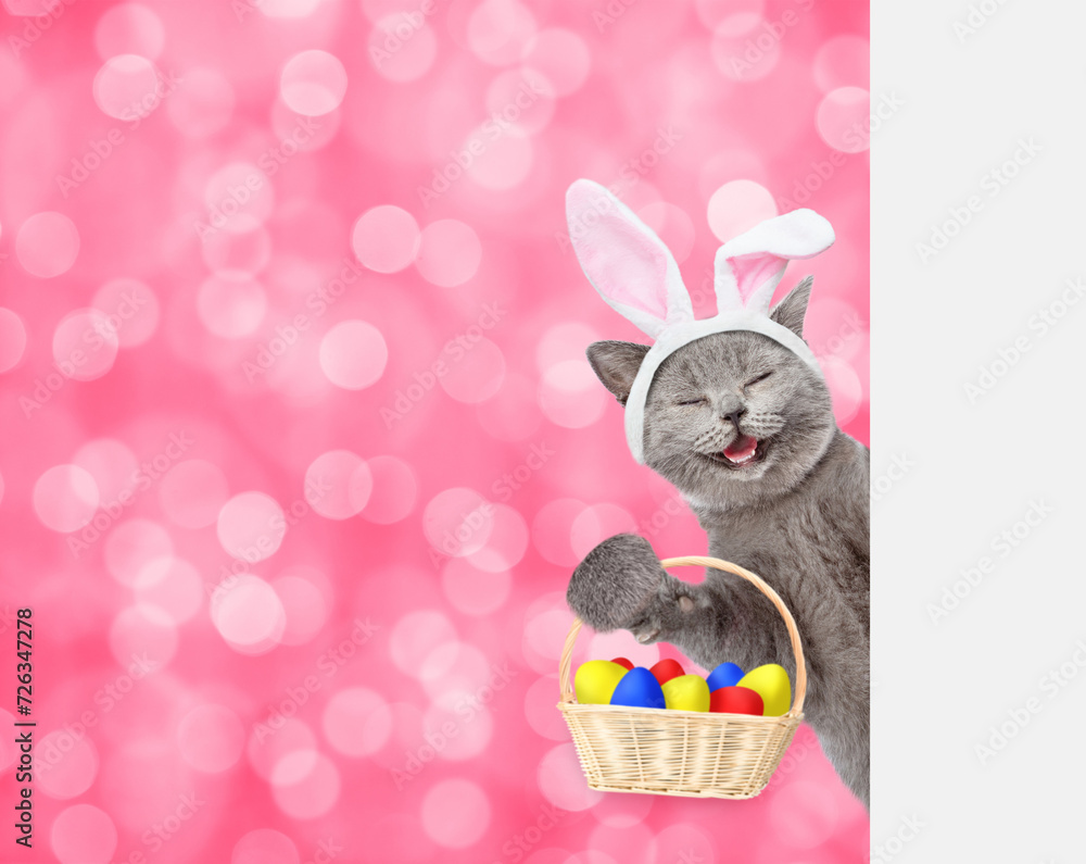 Poster happy kitten wearing easter rabbits ears holds basket with colorful easter eggs and looks from behin