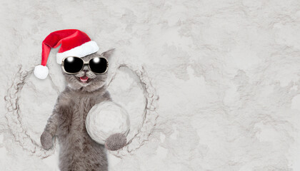 Happy cat wearing sunglasses and red santa hat and scraf holds big snowball while lying on snow....