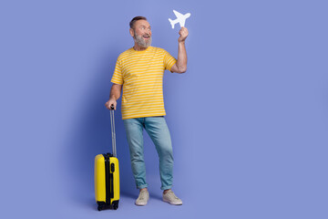 Full size portrait of positive aged person hold suitcase paper plane look empty space isolated on purple color background