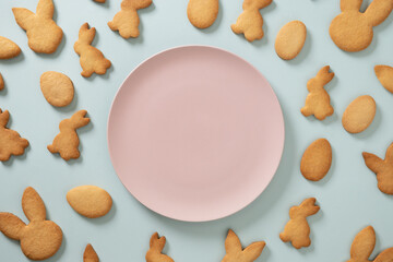Easter cookies shaped of bunny and eggs as pattern and pink plate with space for text on blue background. View from above. Greeting card.