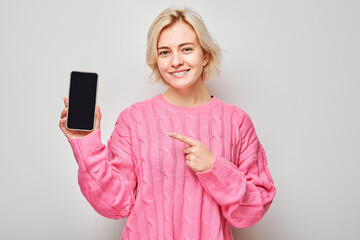 Portrait of young blond woman in pink sweater points finger at mobile phone screen with excited...