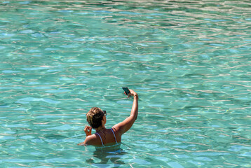 Back view of female tourist taking a selfie in turquoise water during summer vacation