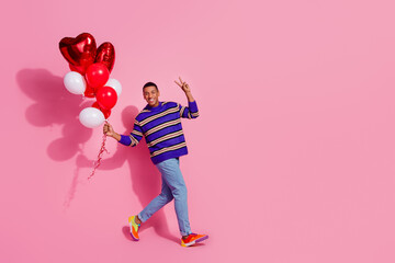 Full length photo of funky cool man wear striped sweater walking showing v-sign rising balloons empty space isolated pink color background