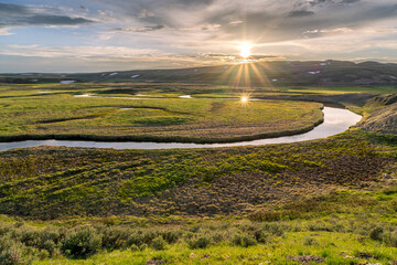 A river meander through a green open meadow with the low sun reflecting off the water at sunset,...