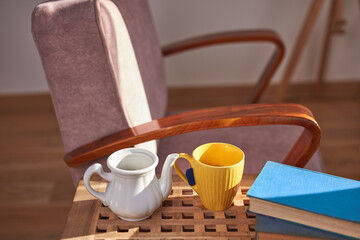 Fototapeta na wymiar Teapot and tea cup with books and a comfortable chair in a sunny home interior.