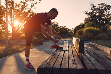 Sporty man tying laces on jogging sneakers in a park. - obrazy, fototapety, plakaty